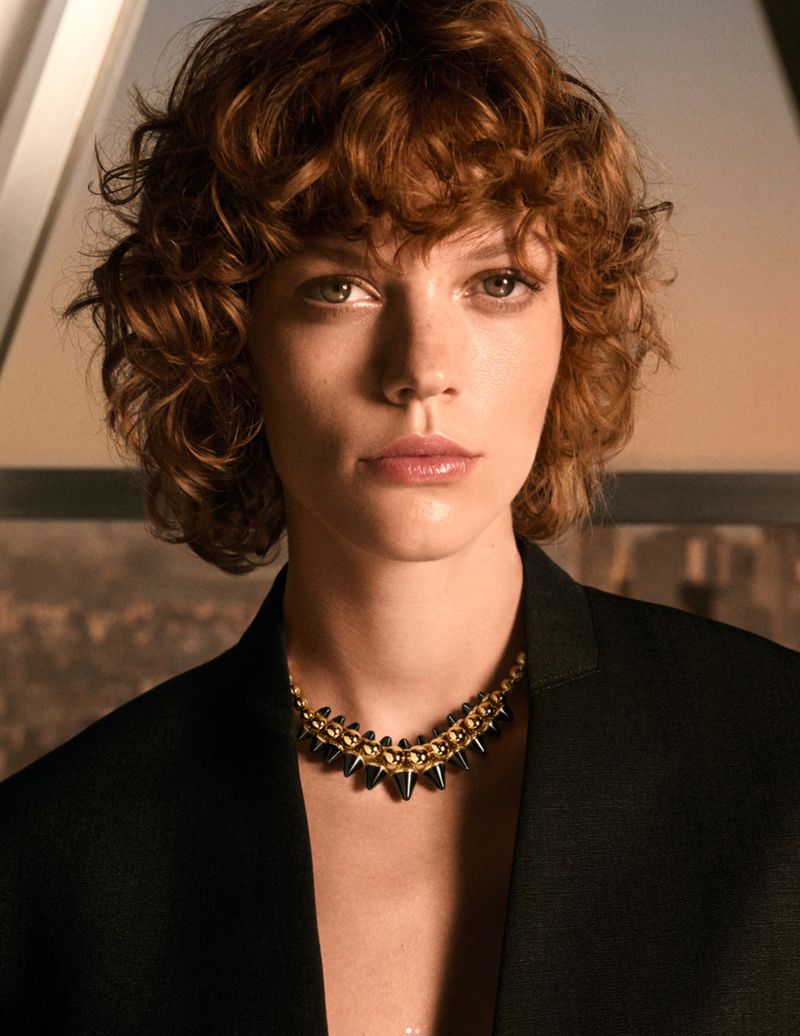 Close-up of a young woman with auburn, curly hair wearing a black jacket and an ornate gold and black necklace, against a city backdrop.