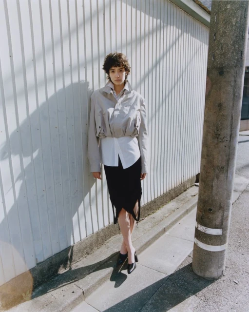Woman in stylish beige jacket and black skirt posing against a corrugated wall on an urban street with harsh sunlight and shadows.