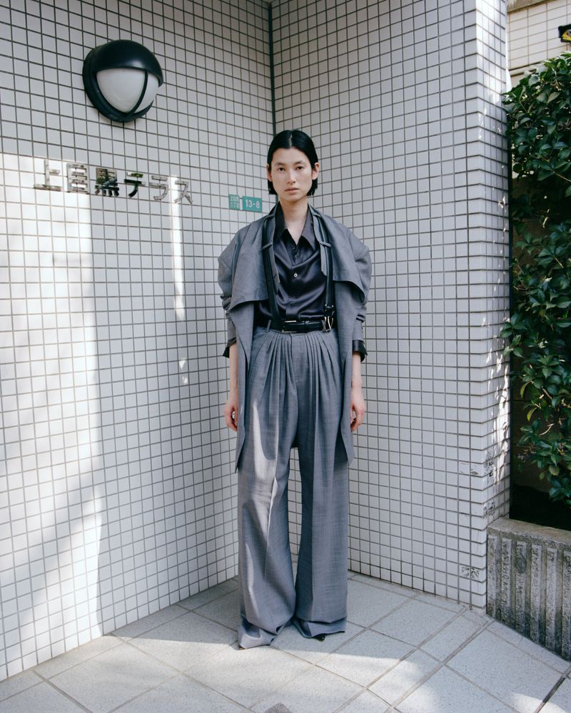 A full-length shot of an Asian woman in a grey oversized suit posing in front of a tiled wall outdoors, with a neutral expression.
