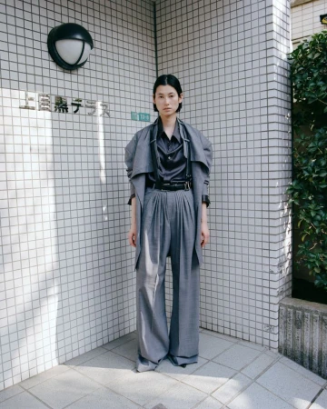 A full-length shot of an Asian woman in a grey oversized suit posing in front of a tiled wall outdoors, with a neutral expression.