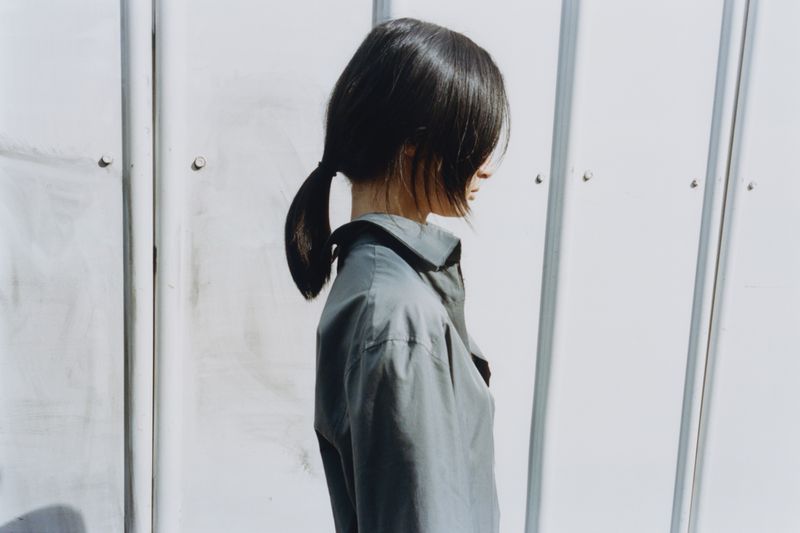 Woman in profile poses in front of a white metal siding wall, wearing a gray button-down shirt with her dark hair in a low ponytail.