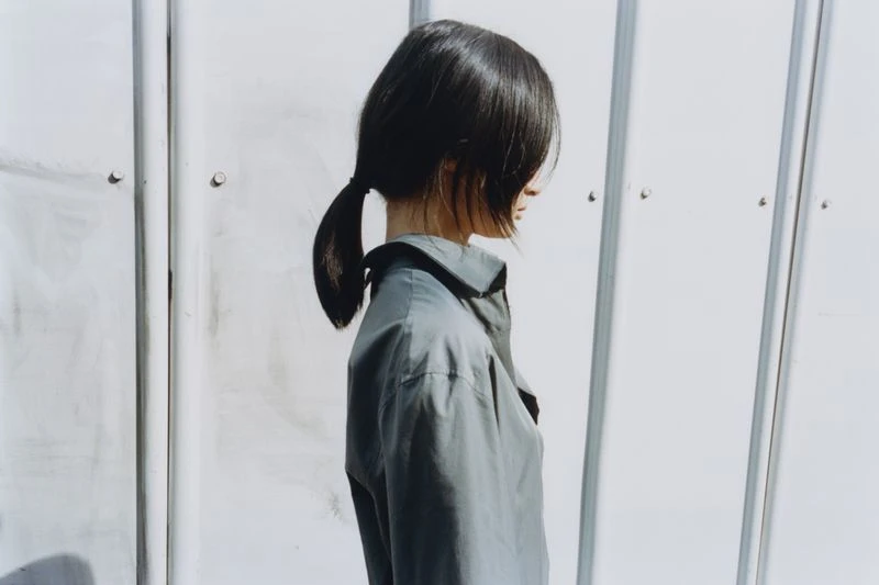 Woman in profile poses in front of a white metal siding wall, wearing a gray button-down shirt with her dark hair in a low ponytail.