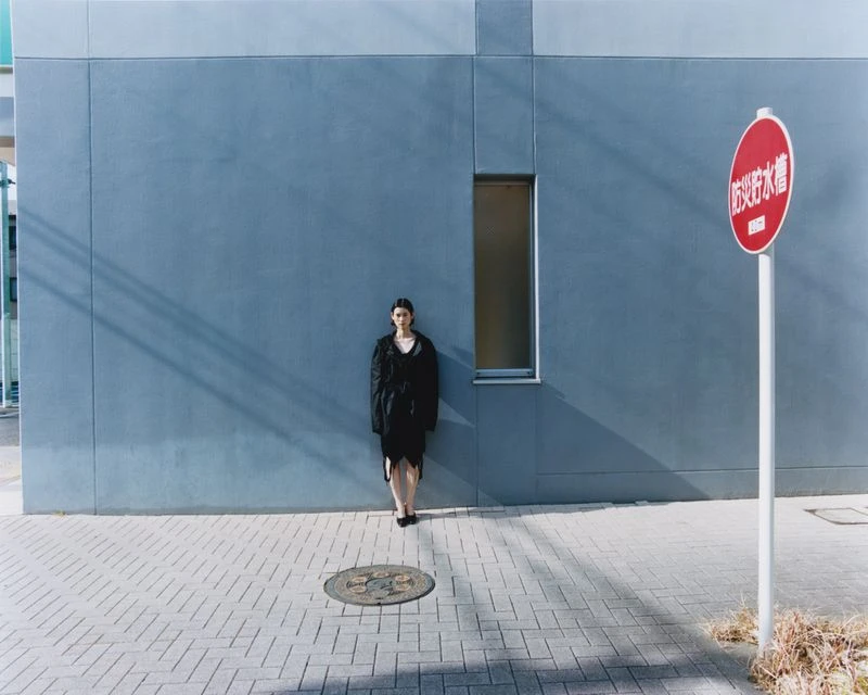 An Asian woman in a black dress stands near a blue building on a sunny day. A red sign and a brick walkway are also visible.