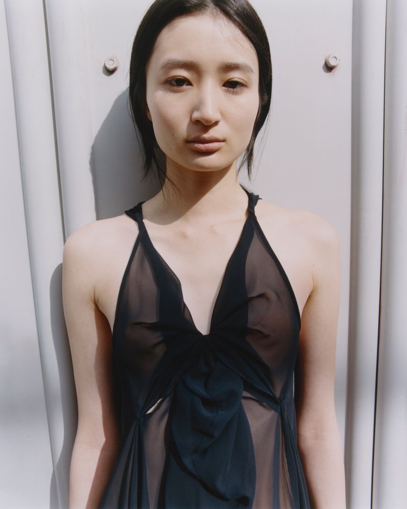 Close-up portrait of a woman with dark hair wearing a sheer black sleeveless top standing against a pale grey industrial wall.