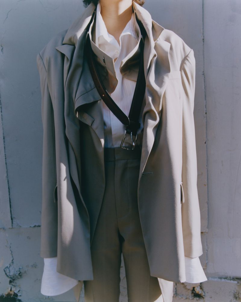 Person in neutral-toned fashion blazer, dress shirt, trousers, and suspenders against a concrete wall in an outdoor environment.