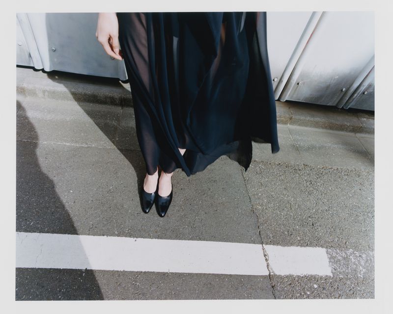 Fashionable adult in a black dress and black leather high heels stands on a cracked concrete surface in an outdoor urban setting.