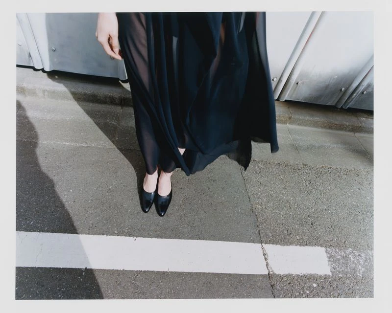 Fashionable adult in a black dress and black leather high heels stands on a cracked concrete surface in an outdoor urban setting.