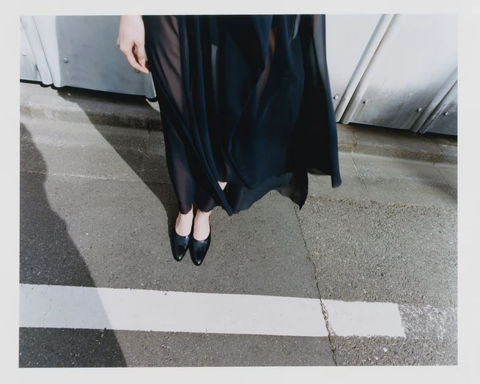 Close-up of a person wearing a sheer dark dress and black shoes standing on pavement next to a white road line.