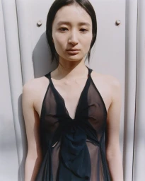 Portrait of a woman wearing a black sheer sleeveless dress standing against a light grey industrial background.