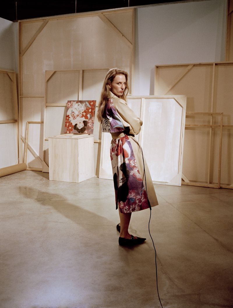Adult woman in a patterned dress poses in a studio setting with a concrete floor and various blank canvases on the wall behind her.
