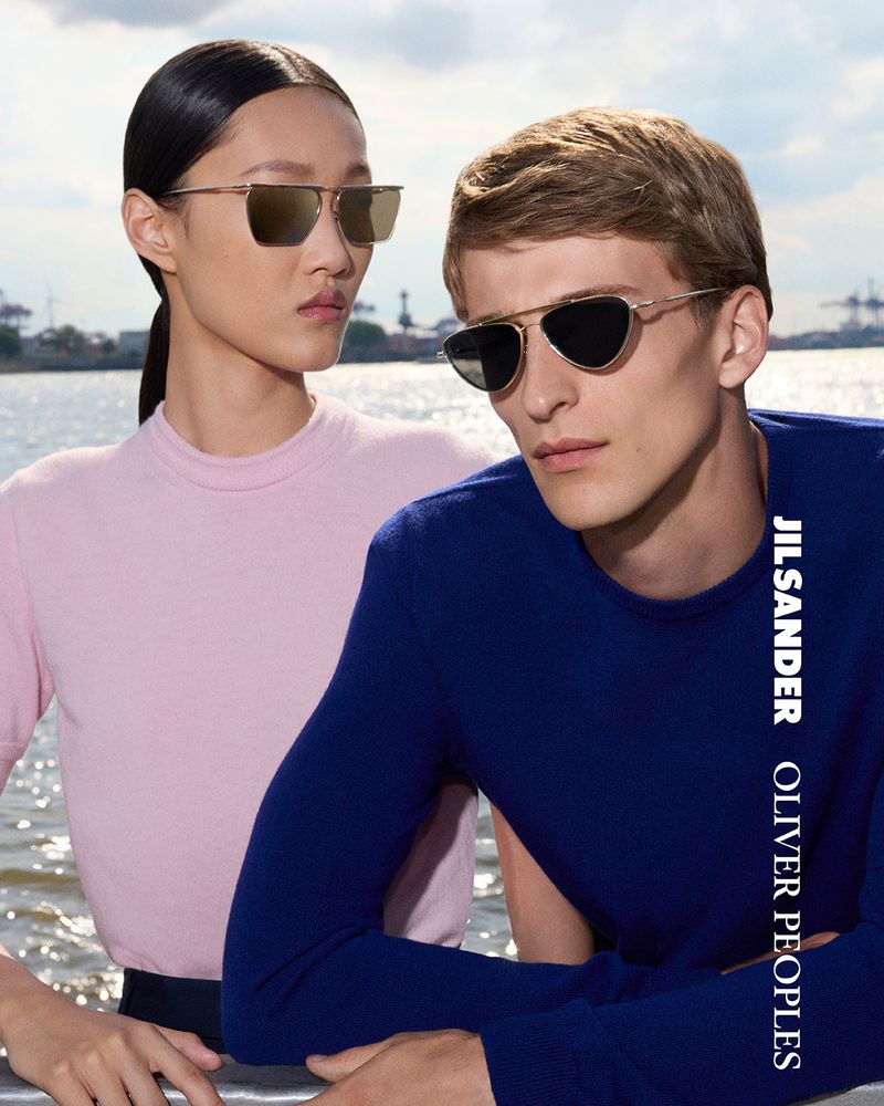 A stylish eyewear advertisement featuring a woman with sunglasses and a man with sunglasses standing in front of a waterfront cityscape.
