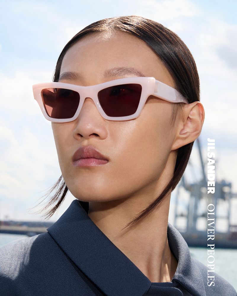 Asian fashion model wearing light pink Jil Sander x Oliver Peoples sunglasses in a close-up shot outdoors with harbor background.