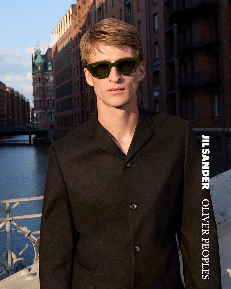 Portrait of a blonde male model wearing green sunglasses and a black suit with a brick building and bridge in the background.