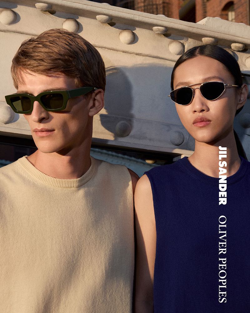 Two fashion models wearing sunglasses and sleeveless shirts pose against a concrete bridge structure on a sunny day in an outdoor setting.