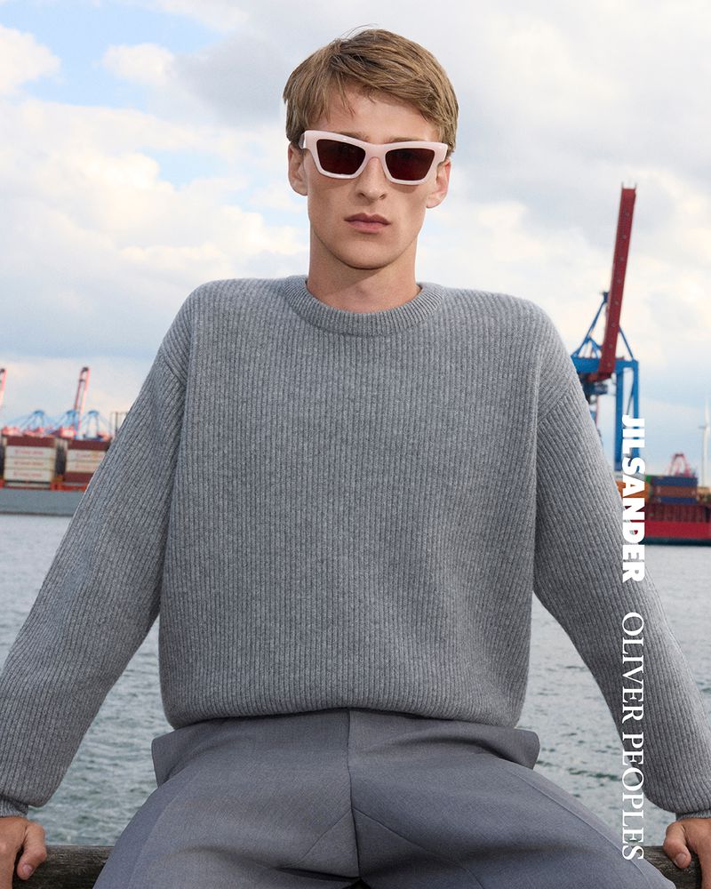 A blond male model poses in a gray sweater and pants with pink sunglasses in front of a harbor with cranes and shipping containers.