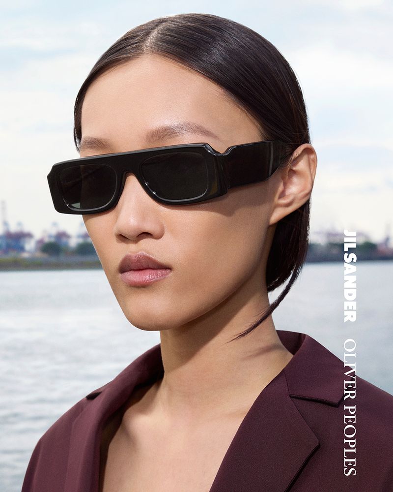A stylish portrait of a person wearing black, thick-framed sunglasses and a maroon blazer with a blurred cityscape background.