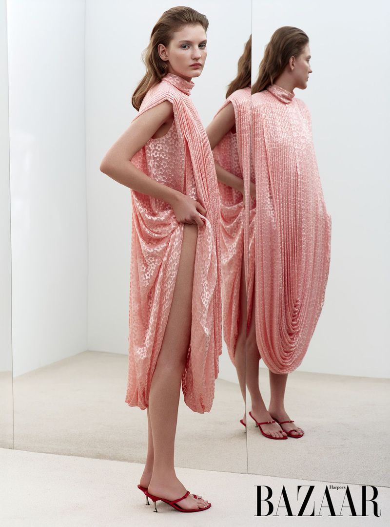 Woman poses in a pink, draped sleeveless dress and red high-heeled sandals, mirrored in a white photo studio against a white background.