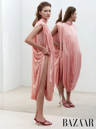 Woman poses in a pink, draped sleeveless dress and red high-heeled sandals, mirrored in a white photo studio against a white background.