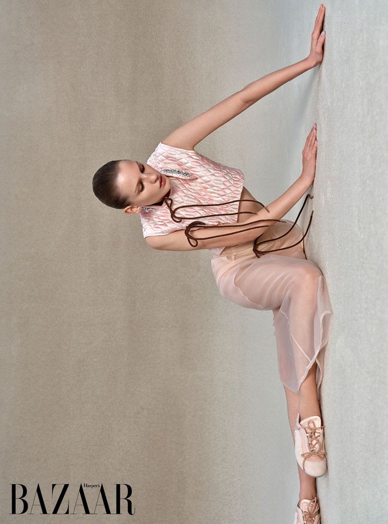 A fashion shot of a young adult woman in a pink and white patterned top and a sheer skirt leaning against a light gray wall.