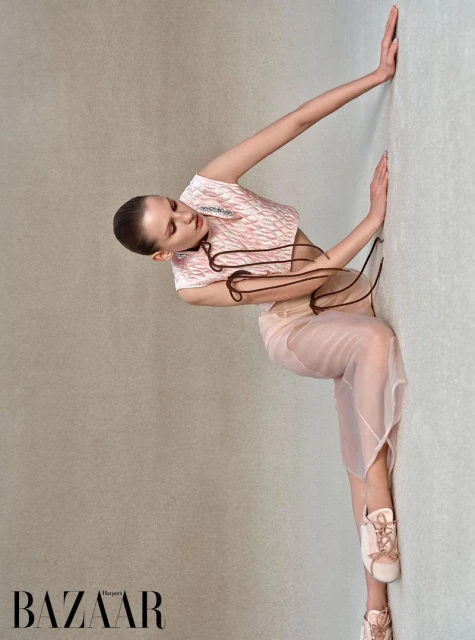 A fashion shot of a young adult woman in a pink and white patterned top and a sheer skirt leaning against a light gray wall.