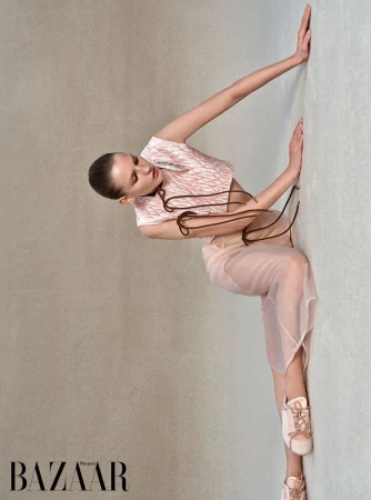 A fashion shot of a young adult woman in a pink and white patterned top and a sheer skirt leaning against a light gray wall.