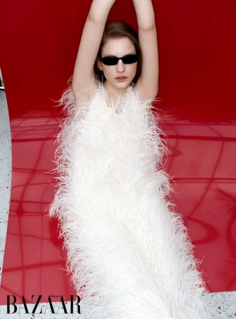 A woman wearing a white feather dress and black sunglasses lies on a reflective red surface with her arms raised above her head.