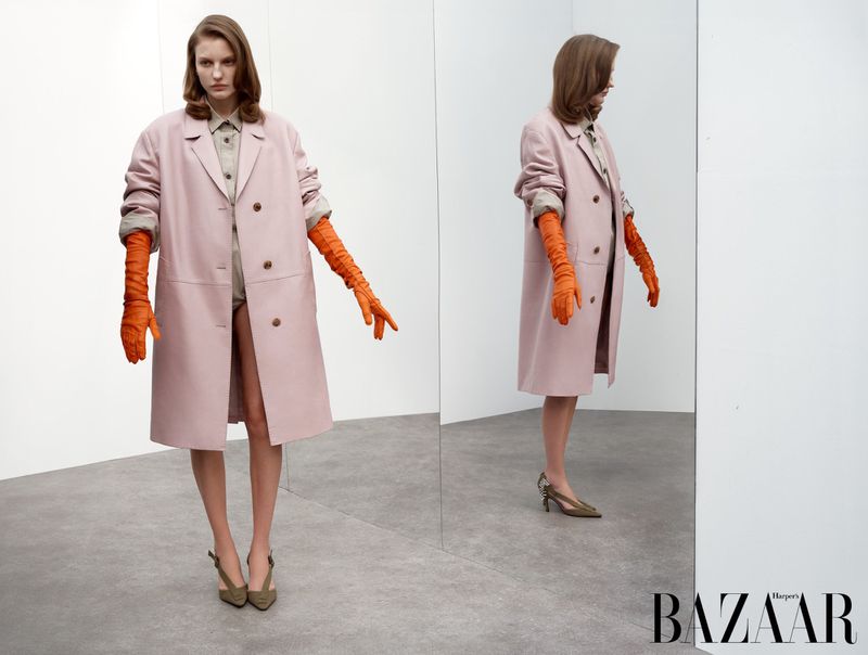 Fashion shot of a woman in a pink coat and orange gloves standing in front of a mirror in a photo studio, Harper's Bazaar logo at the bottom.