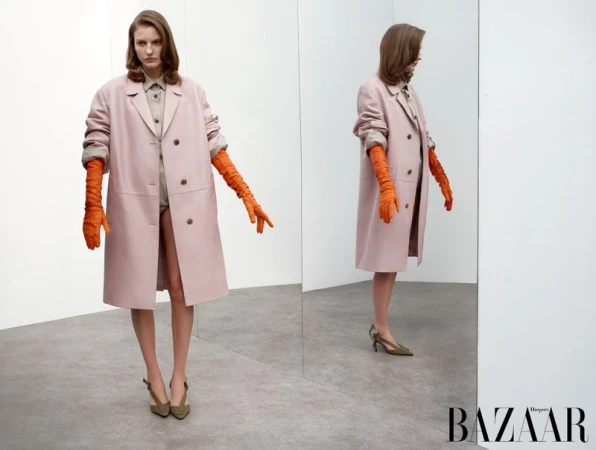 Fashion shot of a woman in a pink coat and orange gloves standing in front of a mirror in a photo studio, Harper's Bazaar logo at the bottom.
