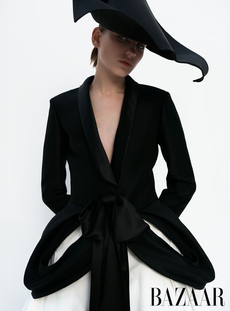 Fashion shot of a woman wearing a black jacket with bow detail and a large black hat against a white backdrop in a photo studio.