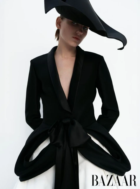 Fashion shot of a woman wearing a black jacket with bow detail and a large black hat against a white backdrop in a photo studio.