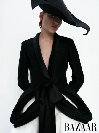 Fashion shot of a woman wearing a black jacket with bow detail and a large black hat against a white backdrop in a photo studio.