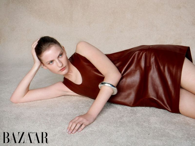 Elegant woman in a brown dress poses on a beige rug in a studio setting, showcasing fashion and style, with a 'Harper's Bazaar' logo visible.