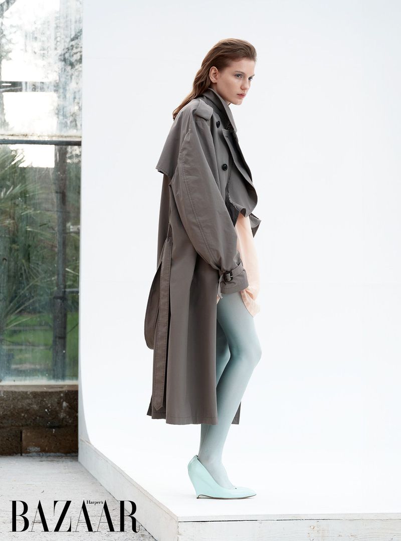 A fashionable woman in a grey trench coat and light blue tights poses in an indoor setting with natural light and a windowed wall.