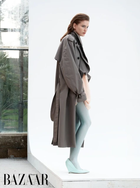 A fashionable woman in a grey trench coat and light blue tights poses in an indoor setting with natural light and a windowed wall.