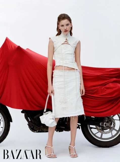 Fashion editorial shot of a female model wearing a white sleeveless top and skirt, holding a white handbag, with a motorcycle.