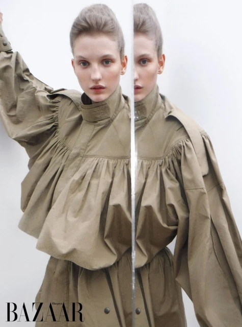 Fashion editorial shot of a woman in a khaki-colored dress with a high collar and voluminous sleeves, reflected in a mirror.