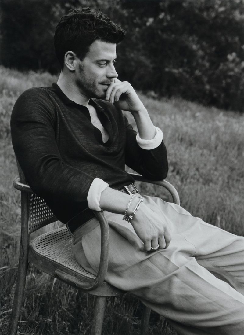 Portrait of adult male seated in chair outdoors, wearing casual shirt and pants in black and white, trees blurred in background.