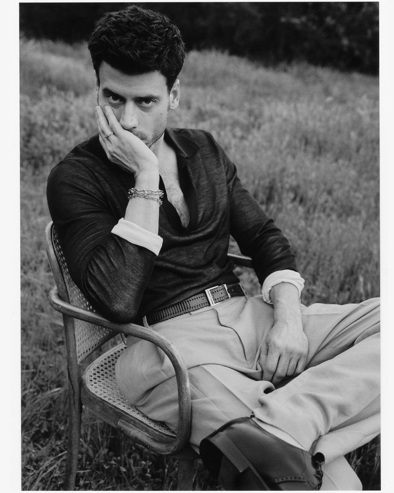 Black and white portrait of a man in a sweater and pants sitting in a chair outdoors in a field of grass.