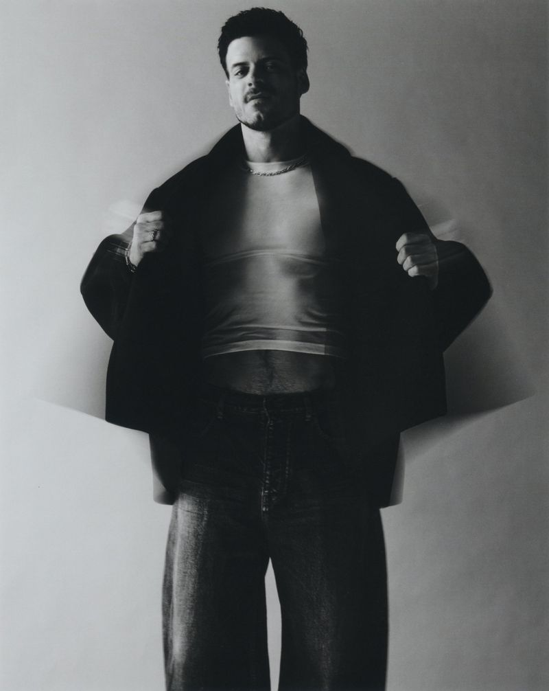 Monochrome portrait of a man with dark hair and mustache in an open black jacket over a striped tee shirt, posing in a photo studio.