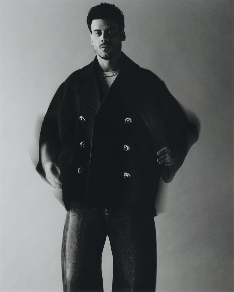 A stylish adult male poses in an oversized black coat and jeans against a neutral background in a studio setting with soft lighting.