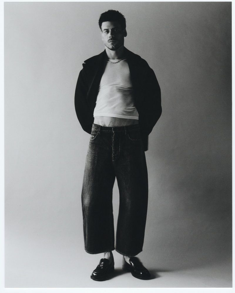Monochrome portrait of a male model with curly hair in an open dark jacket, white t-shirt, wide leg jeans, and dark loafers in a studio.