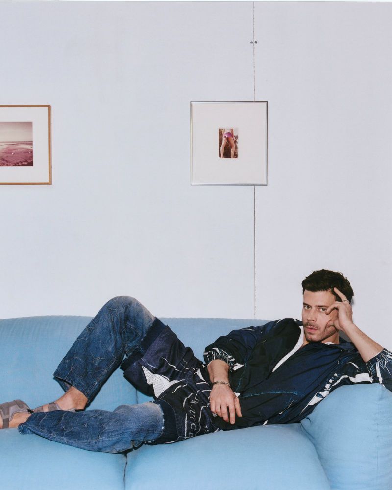 Adult male lounging on a blue couch in a jacket and jeans, indoor setting with artwork on the wall, casual and relaxed pose.