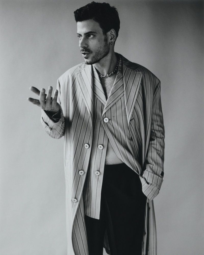 Adult male poses indoors wearing a striped coat and chain. He has dark hair and a beard, in a close-up shot on a neutral background.