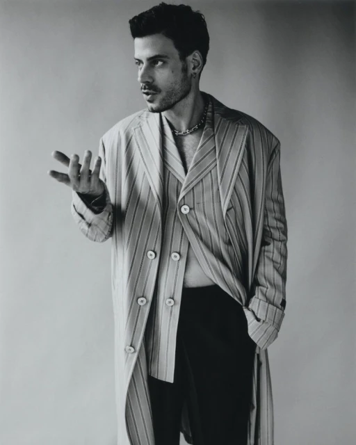 Adult male poses indoors wearing a striped coat and chain. He has dark hair and a beard, in a close-up shot on a neutral background.