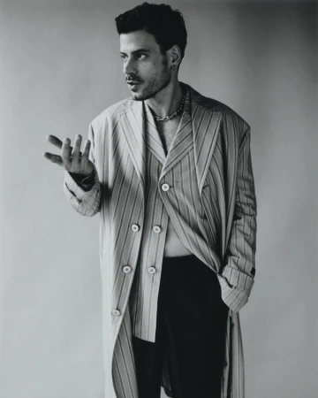 Adult male poses indoors wearing a striped coat and chain. He has dark hair and a beard, in a close-up shot on a neutral background.