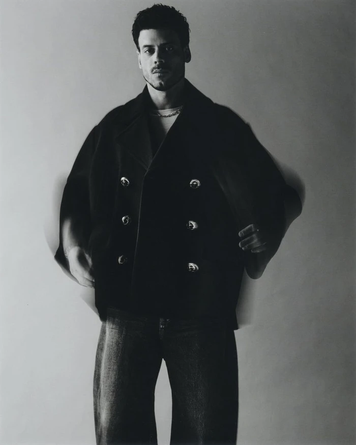Man wearing a black double-breasted jacket and dark trousers against a plain studio background with motion blur on arms