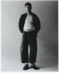Monochrome portrait of a young man wearing a white tank top, dark jacket, and wide-leg jeans against a grey studio backdrop.