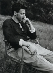 Monochrome portrait of a man seated in a wicker chair outdoors wearing a dark long-sleeve shirt and light trousers.