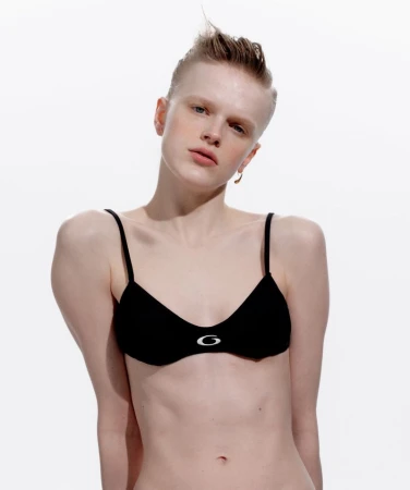 Woman in black spaghetti-strap bra with shaved head in front of white background. Fashion shoot shows her upper body and plain background.