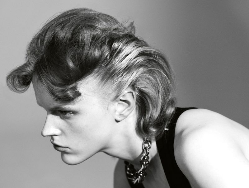 Monochrome portrait of a blonde adult male looking down with curled hair, wearing a black top and a chunky silver necklace.